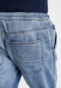 Jeans de mezclilla azul claro con cinturilla elástica, que cuentan con dos bolsillos traseros y una pequeña etiqueta de logo en la parte trasera cerca del bolsillo derecho.