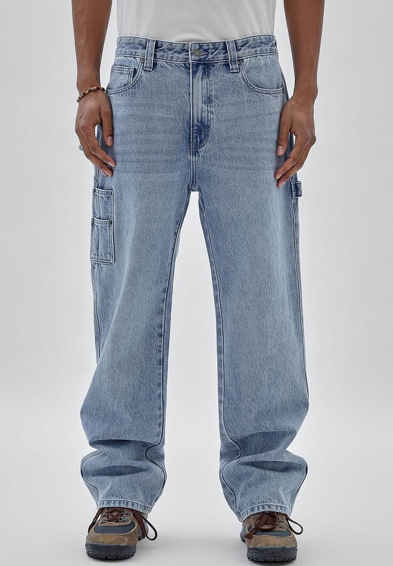 Man draagt losse lichtblauwe cargobroek en bruine wandelschoenen, staand tegen een effen grijze achtergrond.