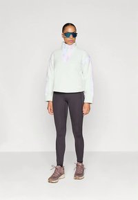 Pullover in pile verde chiaro con cerniera a un quarto e maniche testurizzate; abbinato a leggings neri e scarpe da ginnastica marroni. Indossati occhiali da sole scuri.