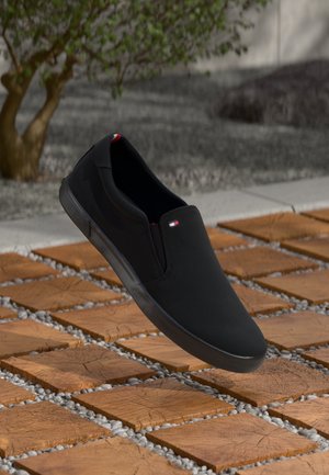 Chaussure noire sans lacets avec un petit logo sur le dessus, flottant au-dessus de carreaux carrés en bois avec des espaces remplis de gravier, à côté d'un arbre en extérieur.