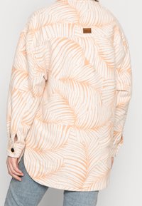 Camicia di flanella con una base bianca e motivo di foglie di palma arancioni, con un orlo posteriore lungo e un'etichetta con il logo Roxy sulla parte superiore della schiena.