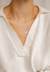 Silberne Kettenhalskette mit einem Halbmondanhänger, getragen über einem weißen, strukturierten V-Ausschnitt-Pullover. Nahaufnahme des Schmucks.