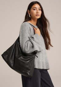 Melna ādas hobu soma ar maigu, teksturētu apdari un savilktu augšu. Modelis valkā pelēku adītu džemperi uz neitrāla fona.