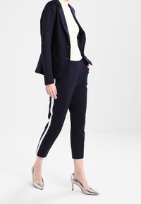Granatowa marynarka z dwoma kieszeniami z przodu, noszona na kremowej bluzce, połączona z granatowymi spodniami z białymi paskami bocznymi oraz srebrnymi szpilkami.