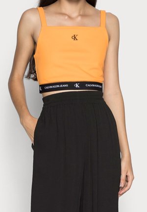 Vrouw draagt een oranje Calvin Klein crop top met dunne bandjes en zwarte hoogtailored broek, hand in de zak rustend.