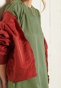 Grünes Kleid mit Rundhalsausschnitt, kombiniert mit einer roten Bomberjacke. Die Jacke hat einen Reißverschluss und gerippte Bündchen und Saum. Glatte Stoffstruktur.