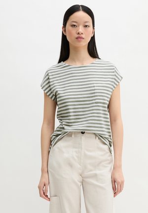 Kvinde med glat sort hår iført en kortærmet grå- og hvidstribet skjorte og beige højtaljede bukser, stående foran en ensfarvet baggrund.