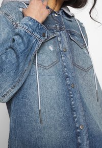 Veste en denim de couleur bleu clair, texture usée, avec des boutons-pression et une capuche grise dotée de cordons. Deux poches poitrine visibles.