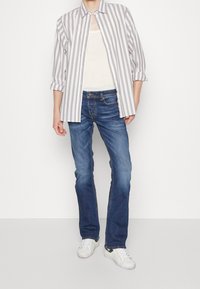 Camisa de botones a rayas en gris y blanco, llevada sobre una camiseta de tirantes blanca. Jeans de mezclilla azul y zapatillas blancas completan el atuendo.