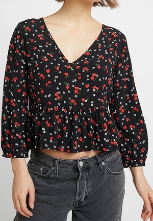 Femme portant un chemisier noir à manches longues en V avec motif floral rouge et blanc et un jean noir délavé taille haute.