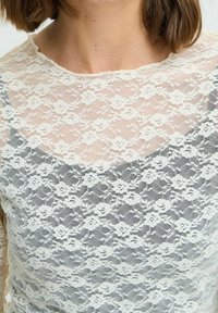 Haut en dentelle avec un motif floral, surmonté d'une superposition ivoire transparente et d'une base unie. L'encolure est ronde et délicatement en dents de scie.
