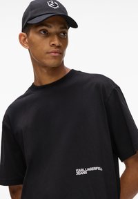 Melns kokvilnas T-krekls ar apaļu kakla izgriezumu, ar baltu "KARL LAGERFELD JEANS" logotipu kreisajā apakšējā stūrī. Valkāts ar melnu cepuri.