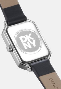 Silberfarbene rechteckige Uhr mit schwarzem Lederarmband und beigem Unterteil, mit einem auffälligen DKNY-Logo auf der Rückseite, wasserdicht bis 3 ATM.