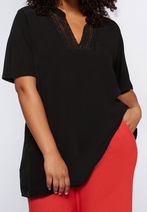 Vrouw draagt een zwart kortermig shirt met kanten V-halsdetail en felrode broek, met één hand in de zak en de andere ontspannen langs haar zij.