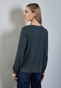 Mörkgrå långärmad topp med rund halsringning och rynkade ärmslut, kombinerad med blå jeans. Slät textur, avslappnad passform, minimal detaljering.