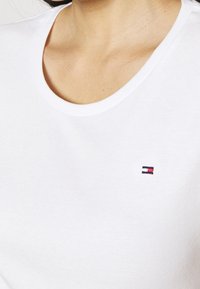 Biała bawełniana koszulka z okrągłym dekoltem i małym haftowanym logo z ciemnoniebieskimi i czerwonymi akcentami w okolicy klatki piersiowej.