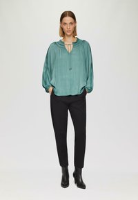 Türkise Bluse mit plissierter Textur, Schlüssel-ausschnitt und gerafften Manschetten, kombiniert mit taillierten schwarzen Hosen und schwarzen ankle boots mit Absatz.