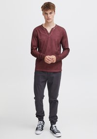 Burgundin pitkähihaisessa henley-paidassa, jossa on napitettava kaula-aukko, yhdistetty tummanharmaisiin jogger-housuihin ja mustiin korkeavyötäröisiin tennareihin, seisoo vaaleaa taustaa vasten.