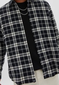 Uomo che indossa una camicia a quadri bianca e nera sbottonata sopra una maglietta nera, accessoriato con due collane a catena d'argento.