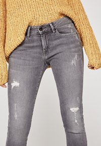 Jeans de mezclilla gris con un corte ajustado, que presentan áreas desgastadas y detalles de efecto arrugado. Combinados con un suéter de punto suelto en mostaza.