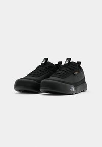 Black athletic shoes made of fabric and rubber, featuring a rounded toe, textured soles, and a pull tab on the heel. Logo displayed on the side.