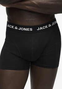 Μαύρα boxer briefs με λευκό λογότυπο "JACK & JONES" στη ζώνη, φορεμένα από άτομο με σκούρο τόνο δέρματος, περικομμένα από τη μέση μέχρι τη μέση του μηρού.