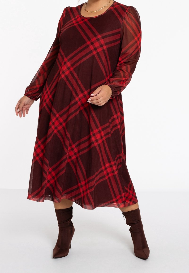 Femme portant une robe à manches longues bordeaux à motif tartan rouge et des bottines marron, posant devant un fond uni.