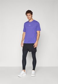 Homme debout portant une chemise à manches courtes violette, un short noir, des leggings noirs et des chaussures de course blanches sur un fond clair uni.