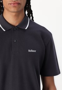 Cămașă polo de culoare bleumarin din material cotone, având un guler clasic, placket cu trei butoane și cusături albe de accent. Logo-ul brodat Hollister.