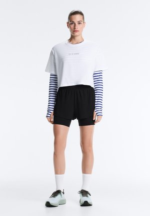 Hölgy, akinek a haja hátra van kötve, fehér rövidített pólót visel kék-fehér csíkos hosszú ujjú felső felett, fekete rövidnadrágot, fehér zoknit és halványkék tornacipőt.