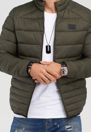 Mężczyzna w oliwkowej pikowanej kurtce, białej koszulce, niebieskich podartych dżinsach, czarnym naszyjniku z zawieszką typu dog tag, skórzanej bransoletce oraz czarnym zegarku ze złotymi akcentami.