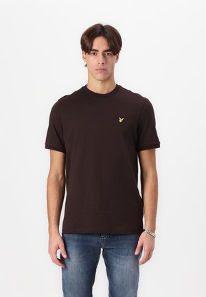 Tricou din bumbac maro, cu mâneci scurte, guler rotund și un logo brodat galben pe partea stângă a pieptului; asortat cu blugi din denim albastru.