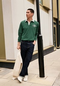 Zelená polokošeľa s dlhým rukávom a bielym golierom, tmavé džínsové nohavice a biele tenisky. Model drží svetlo béžovú mikinu. Městské pozadie.