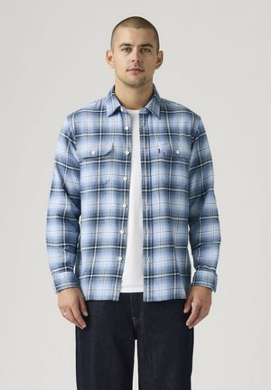 Joven con el pelo corto que lleva una camisa azul y blanca de cuadros abotonada sobre una camiseta blanca y vaqueros oscuros, de pie frente a un fondo liso.