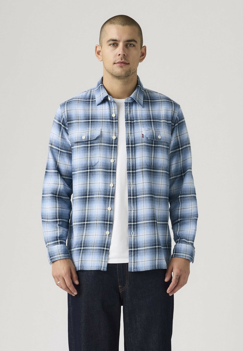 Ung mand med kort hår iført en blå og hvid ternet skjorte med knapper over en hvid t-shirt og mørke jeans, står foran en ensfarvet baggrund.