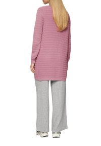 Roze gebreide lange sweater met een textuurdesign, gecombineerd met grijze wijde broeken. De sweater heeft lange mouwen en zijsplitten.