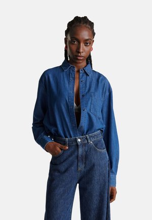 Camisa de mezclilla azul oscuro con un bolsillo, diseño de botones y corte amplio. Combinada con pantalones de mezclilla oscuros de tiro alto.