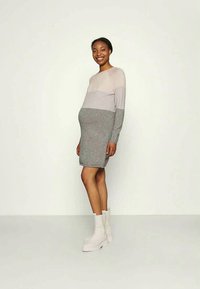 Kršna obleka za nosečnice z barvno blokado v svetlo bežni in sivi barvi. Dolgi rokavi, mehak pleteni material in dolžina do kolen. Bele gležnjarje.