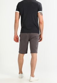 Muž stojící zády, oblečený v černém tričku s kontrastním lemem, šedých šortkách ke kolenům, bílých teniskách a s tetováním na levé ruce.