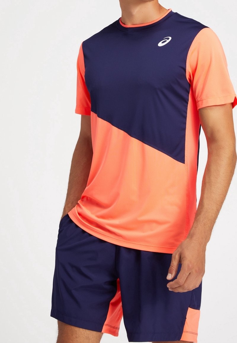 Homme portant un t-shirt de sport à manches courtes bleu marine et corail avec un short bleu marine assorti, debout devant un fond uni et clair.