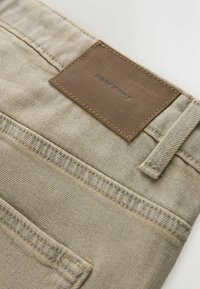 Beige Jeans aus Denim im Nahaufnahme, die die Gesäßtasche, Gürtelschlaufe und ein braunes Lederetikett mit eingeprägtem Schriftzug "DENIM SUPPLY" zeigen.