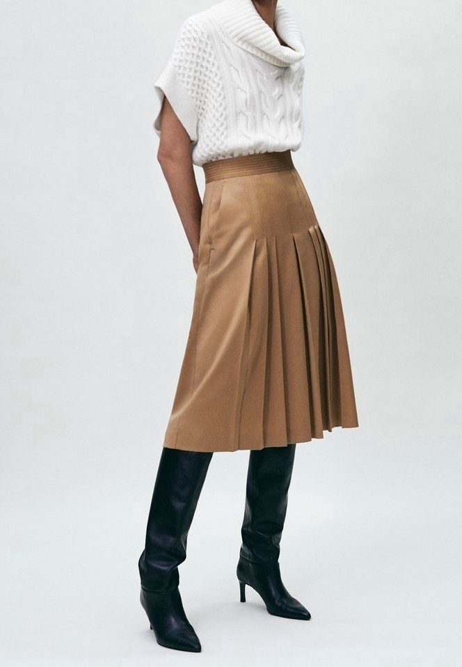 Jupe plissée beige à taille haute, associée à un pull en maille torsadée blanc. Le modèle porte des bottes montantes noires à talons aiguilles.