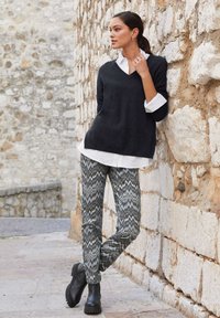 Dunkelgrauer Strickpullover mit V-Ausschnitt, über einem weißen Hemd mit Knöpfen getragen. Graue Hosen mit Zickzackmuster und schwarze Ankle-Boots vervollständigen den Look.