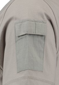 Primer plano de tela gris claro con un bolsillo rectangular con tapa, cosido en la manga de una prenda.