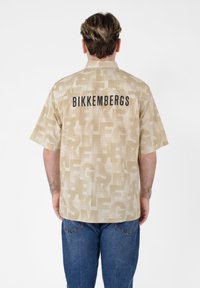 Camicia beige a maniche corte con motivo tonale che presenta il testo "BIKKEMBERGS" in nero sul retro. Il tessuto ha una finitura testurizzata.