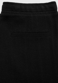 Pantalons en tissu noir texturé avec une taille élastique et une seule poche arrière horizontale sans fermeture visible.