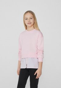 Hellrosa, verkürzter Sweatshirt mit langen Ärmeln, weicher Textur, kombiniert mit einem darunterliegenden gestreiften Hemd in Weiß und Hellblau sowie schwarzen Jeans.
