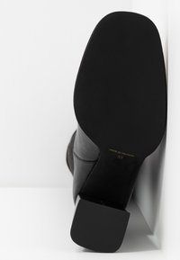 Semelle d'une botte noire à talon bloc, inclinée contre une surface blanche, affichant la taille 37 et l'étiquette "Fabriqué au Vietnam".