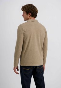 Mann mit lockigem Haar, der zur Seite gedreht ist, trägt ein langärmeliges beiges Poloshirt und dunkelblaue Jeans vor einem schlichten hellen Hintergrund.