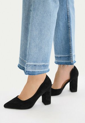 Persona con vaqueros cortos azul claro y zapatos de tacón grueso de ante negro, de pie sobre fondo blanco.